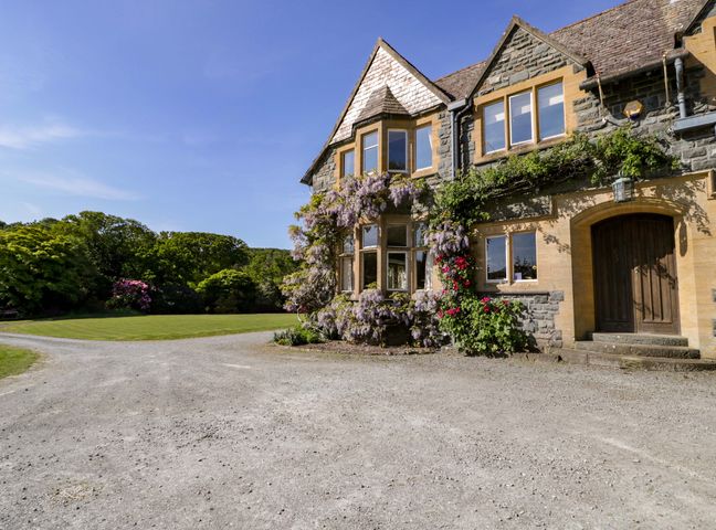 A building with shrubs and flowers by the entrance with a gravel driveway and grassy area at Plas Gwynfryn in Llanbedr