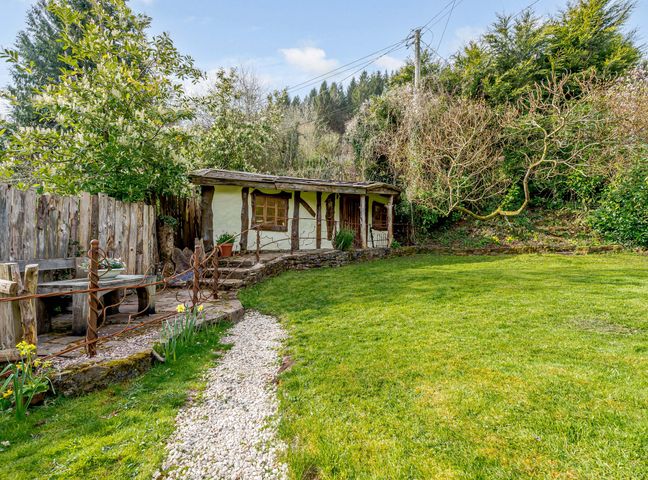 A garden with a grassy lawn a gravel path wooden bench and a small building with timber framing at Brookbank Folly in Mitcheldean