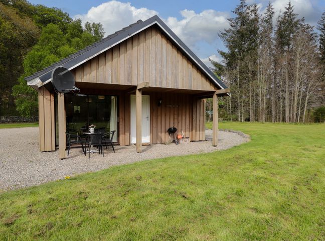 A wooden cottage with an outdoor table and chairs on gravel next to a grassy lawn and trees at Stone Water Cottage in Strathpeffer
