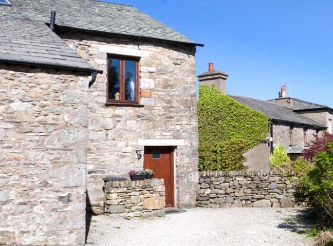 A stone house with wooden door and window surrounded by stone walls and greenery at The Byre in Field Broughton near Cartmel