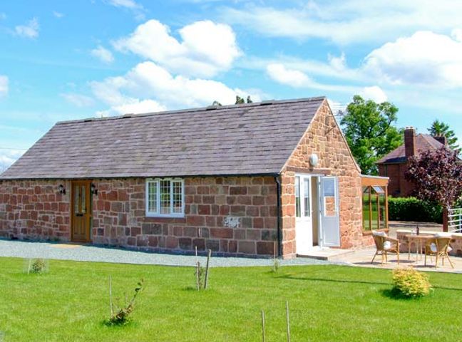A small stone cottage with a tiled roof and wooden door next to a patio with chairs and a table at Byre Cottage Nesscliffe near Shrewsbury