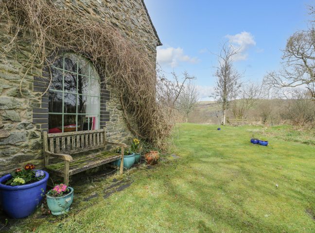 The exterior of a stone building with a wooden bench and flower pots on a grassy lawn at Beudy Dolpebyll in Llangadfan