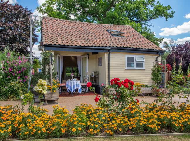 A small cottage with a tiled roof and open double doors leading to a table with two chairs surrounded by flower beds with yellow and red flowers at Norbank Cottage in Bressingham