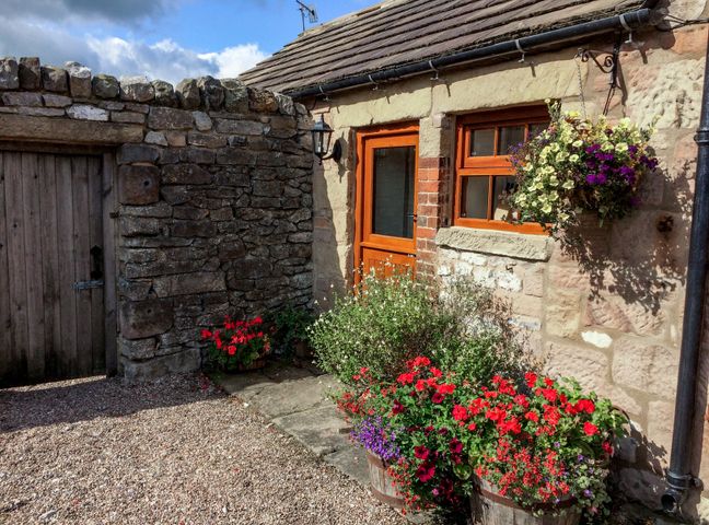 An outdoor area with a stone wall wooden door wooden framed window and door flower pots and hanging basket at The Cow Shed in Alport near Bakewell