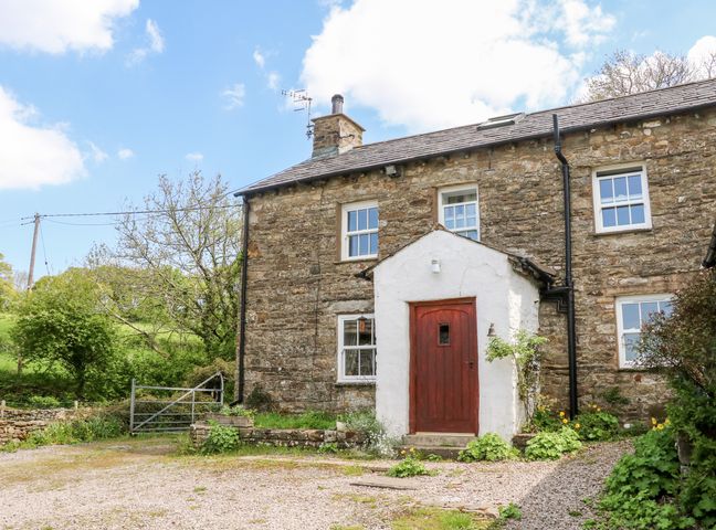 A stone cottage with a red door and white porch surrounded by greenery and gravel at Spout Cottage in Sedbergh