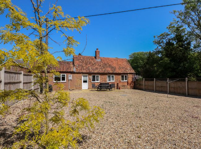 A small brick house with a tiled roof and a gravel yard with outdoor furniture and a tree at Hadleigh Farm Cottage in Gayton