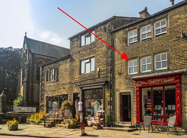 A street view of stone buildings with shops including a sweet shop with a red storefront and outdoor chairs and tables at Church View in Haworth