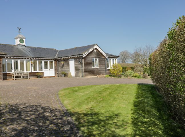 A building with wooden siding and a clock tower with a weathervane surrounded by a paved courtyard and grass lawn with hedges at Clock Tower Cottage in Great Bedwyn near Marlborough