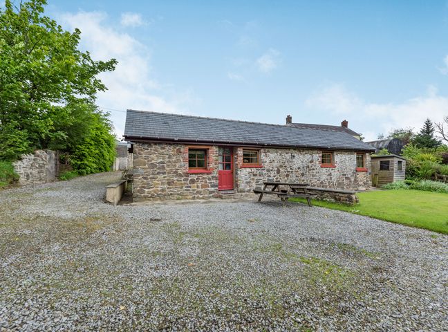 A stone cottage with a red door and windows a picnic table a gravel driveway and a grassy area at Cilwen Fach in Cynwyl Elfed