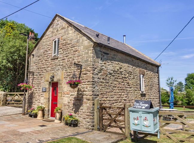 A stone building with a red door and hanging flower baskets next to a wooden gate with a sign for free range eggs at The Old Chapel in Crich