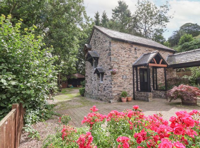 A stone building with a tiled roof next to a paved area and pink flowers at The Old Barn in Llanferres near Ruthin