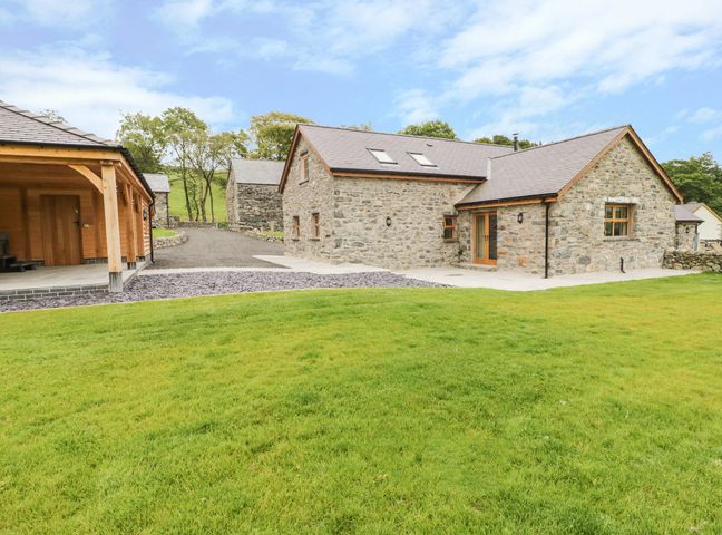 A stone house with wood framed windows and a covered wooden structure on a lawn at Sgubor in Llangwm near Bala