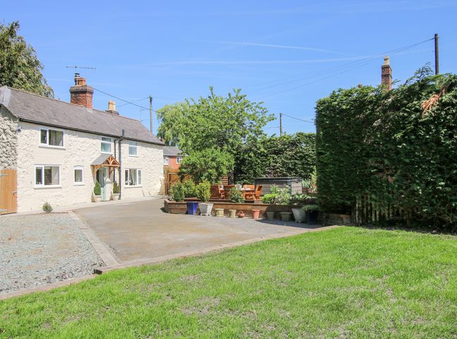 A white cottage with a gravel driveway wooden seating area and hedges at Pennal Cottage in Maesbury Marsh near Knockin