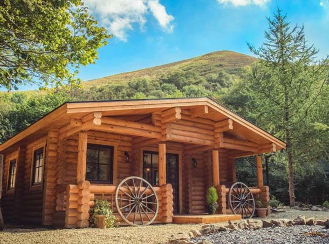A wooden log cabin with wagon wheels on the porch and trees around at Wilderness Lodge in Stiperstones