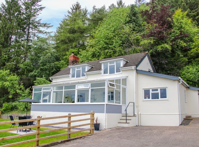 A white house with large windows and a small patio area with a table and umbrella surrounded by trees at Caesamson in Caersws