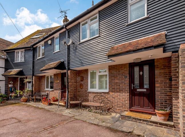 A row of brick and black wooden townhouses with front doors benches and potted plants at Rye House in Rye