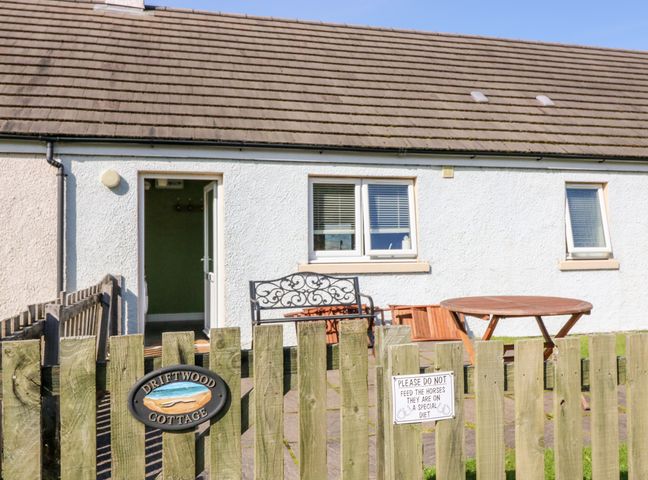 A white cottage with two windows and a door behind a wooden fence with a bench and a round wooden table at Driftwood Cottage Craignure Isle Of Mull