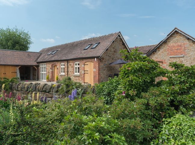 A stone cottage with wooden doors and windows surrounded by green bushes and flowering plants at Swallows Cottage in Harley near Much Wenlock