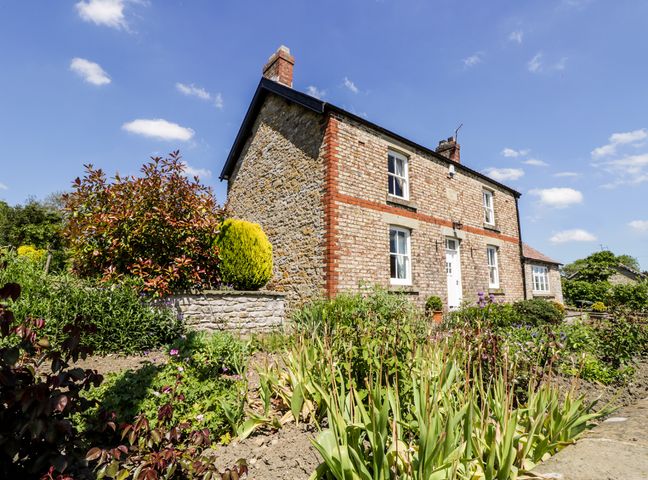 A stone and brick house with a garden area containing various plants and shrubs under a blue sky at Auburn House in Beadlam near Helmsley