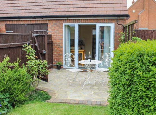 A patio with a white metal table and two chairs with a bottle and glasses near a brick building with open white double doors and surrounding greenery at The Herb Garden in Great Carlton