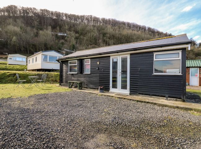 A black wooden chalet with white framed windows and doors on a gravel and grass area with outdoor chairs near trees and mobile homes at Clarach Bay Holiday Village near Aberystwyth
