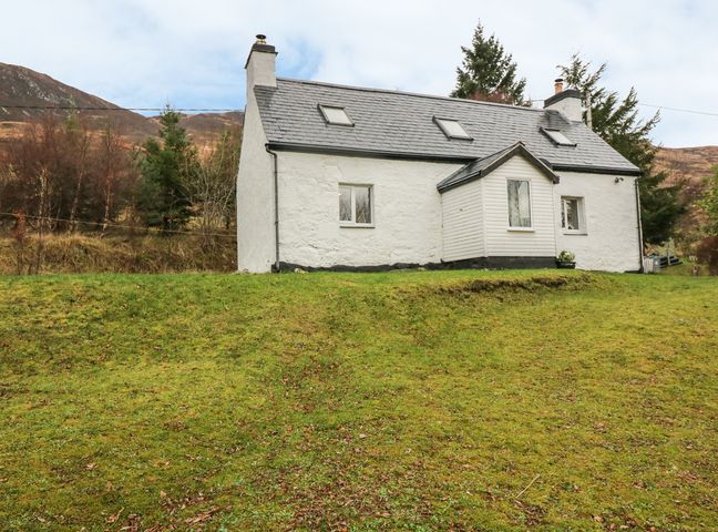 A white cottage with a sloped roof and skylights on a grassy hill with trees and hills in the background at Creag Mhor Cottage in Dornie