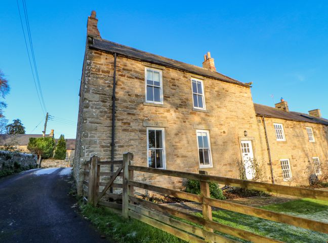 A stone farmhouse with four windows and a wooden fence along a narrow road at East Farmhouse Cottage in Humshaugh near Hexham