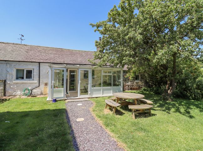 A garden with a round wooden picnic table and benches under a tree by a house with a glass porch at Dyffryn Bach in Glan-yr-afon near Corwen
