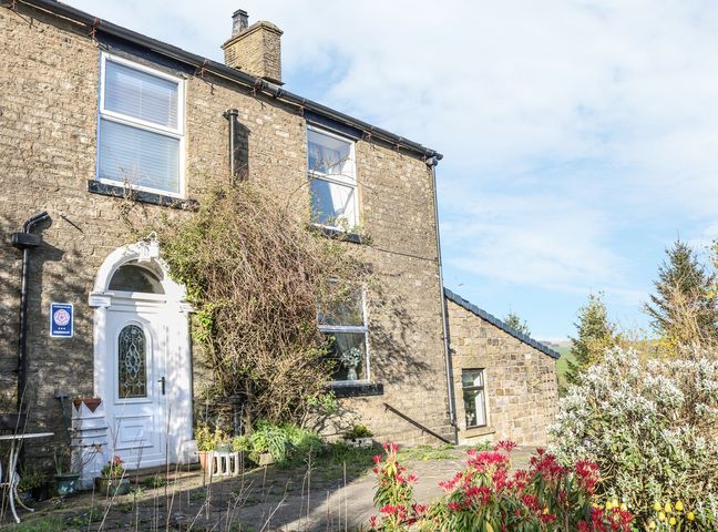 The exterior of a stone house with a white door and windows surrounded by plants and bushes at The Cottage at Moseley House Farm in Chinley