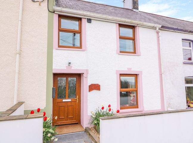 The front of a two-story house with a wooden door and brown framed windows with red and white flowers planted outside at Bank House in Capel Iwan near Newcastle Emlyn