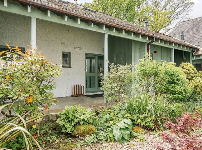 An exterior view of a building entrance with green door and wooden paneling surrounded by various bushes and plants at Cherry - Woodland Cottages in Bowness-in-Windermere