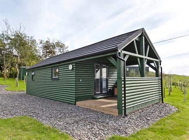 A green wooden cabin with a covered porch and gravel pathway at Ash Lodge in Pennington near Ulverston