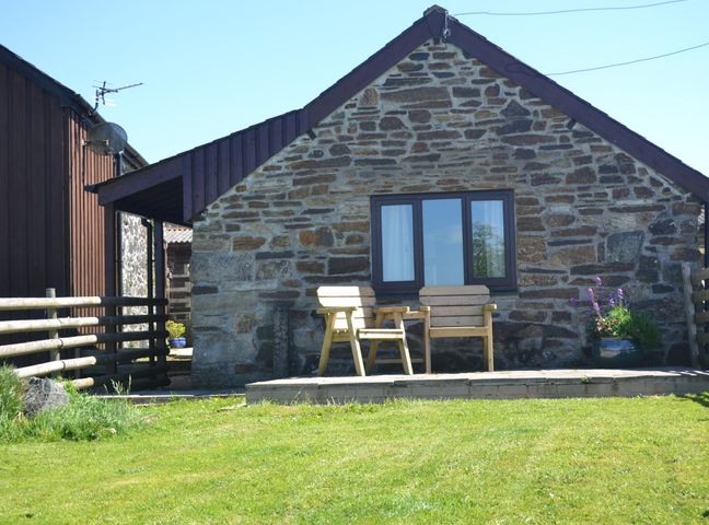 A stone cottage with two wooden chairs and a small table on a patio with a grassy lawn at Nestling Barn Peter Tavy near Tavistock