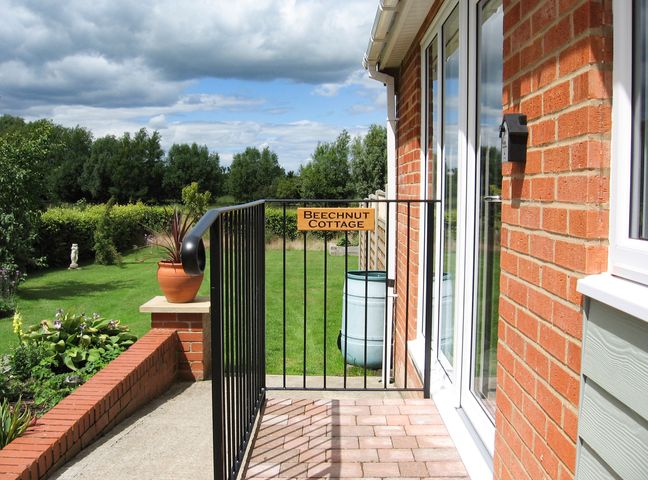 A side entrance to a brick house with a black metal gate with a Beechnt Cottage sign and a garden with green grass and trees in the background at Beechnut Cottage in Tewkesbury