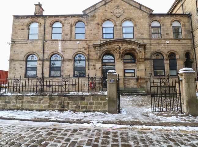 A stone building with arched windows and an arched entrance behind a black iron fence with a gate on a snowy cobblestone street at Apartment 24 in Keighley