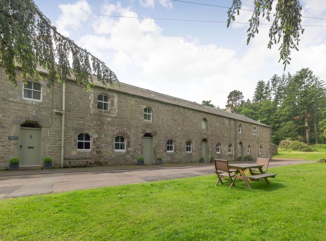 An outdoor seating area with a wooden table and benches on grass in front of a long stone building surrounded by trees at Gardeners Cottage in Bellingham