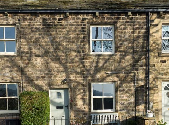 A stone house facade with four windows a pale green door a bench and a hedge at Beacon Cottage in Addingham