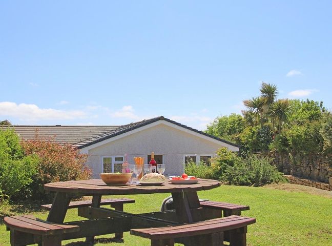 A wooden picnic table with food and drinks in a garden with a house and plants at Carne Vue in Penzance