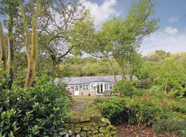 A stone cottage surrounded by trees and shrubs with a picnic bench in front at Dairy Cottage in Bodmin