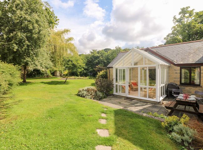 A garden area with green grass trees and a glass conservatory attached to a stone house at Tre Pol Pen in Falmouth