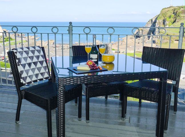 A balcony with a glass table and four chairs overlooking the sea with a bottle, glasses of orange juice, and fruit at 5 Arlington Villas in Ilfracombe