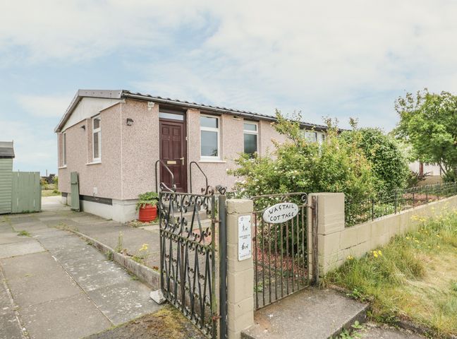 Exterior view of a single-story house with a dark wooden door a metal gate with a sign reading Wagtail Cottage and a garden in Southerness