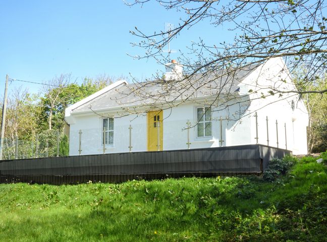 A white cottage with a yellow door and glass railing surrounded by grass and trees at Hidden Gem Cottage in Lettermacaward near Dungloe County Donegal