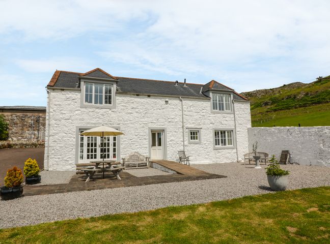 A white stone cottage with outdoor benches a picnic table with an umbrella and potted plants at Bracken Holiday Cottage in Dalbeattie