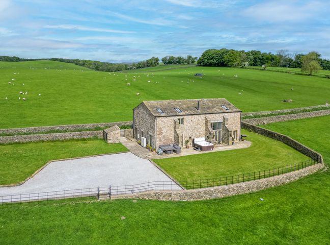 A stone house with a patio and hot tub surrounded by green fields and stone walls at Crane Field Laithe in Otterburn North Yorkshire