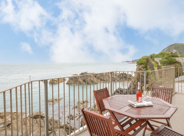 A balcony with a wooden table and chairs overlooking rocky coastline and sea at Oceans Side in Ilfracombe