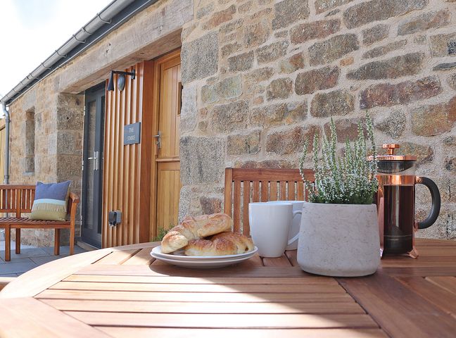 A wooden outdoor table with croissants on a plate two white mugs a small potted plant and a coffee press on the patio outside a stone building at The Stable Boskensoe Barns in Mawnan Smith near Falmouth
