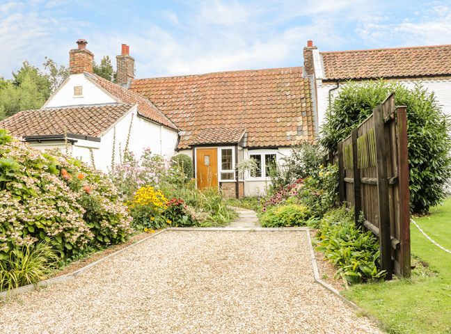 A cottage with a tiled roof surrounded by flowers and plants with a gravel path and a wooden fence at Mrs Dales Cottage in Clenchwarton near Kings Lynn