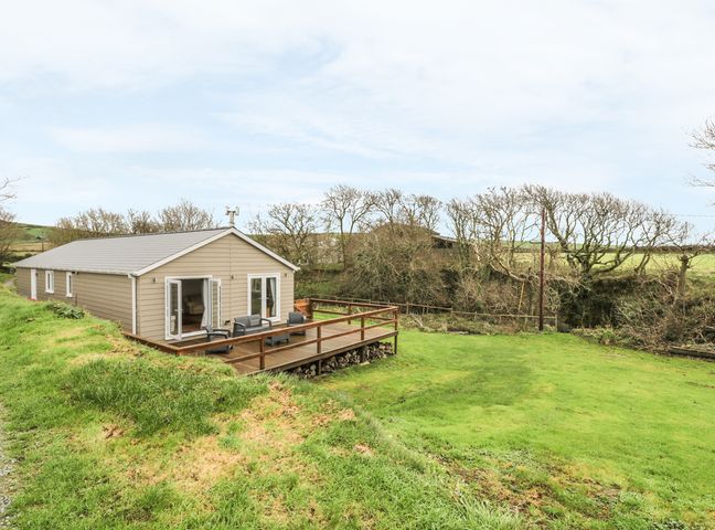 A single-story building with beige siding and a wooden deck on a grassy area with trees in the background at Nant Y Felin Lodge in Aberdaron