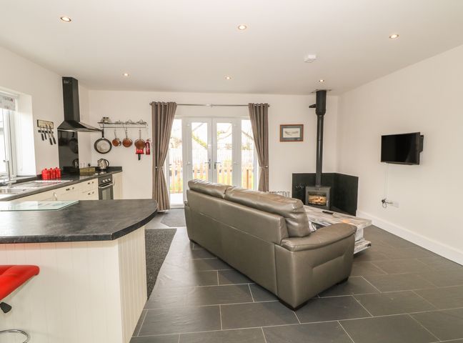 An open plan kitchen and living room with a gray sofa a wood stove and wall mounted television at Hafod Wen in Pentraeth